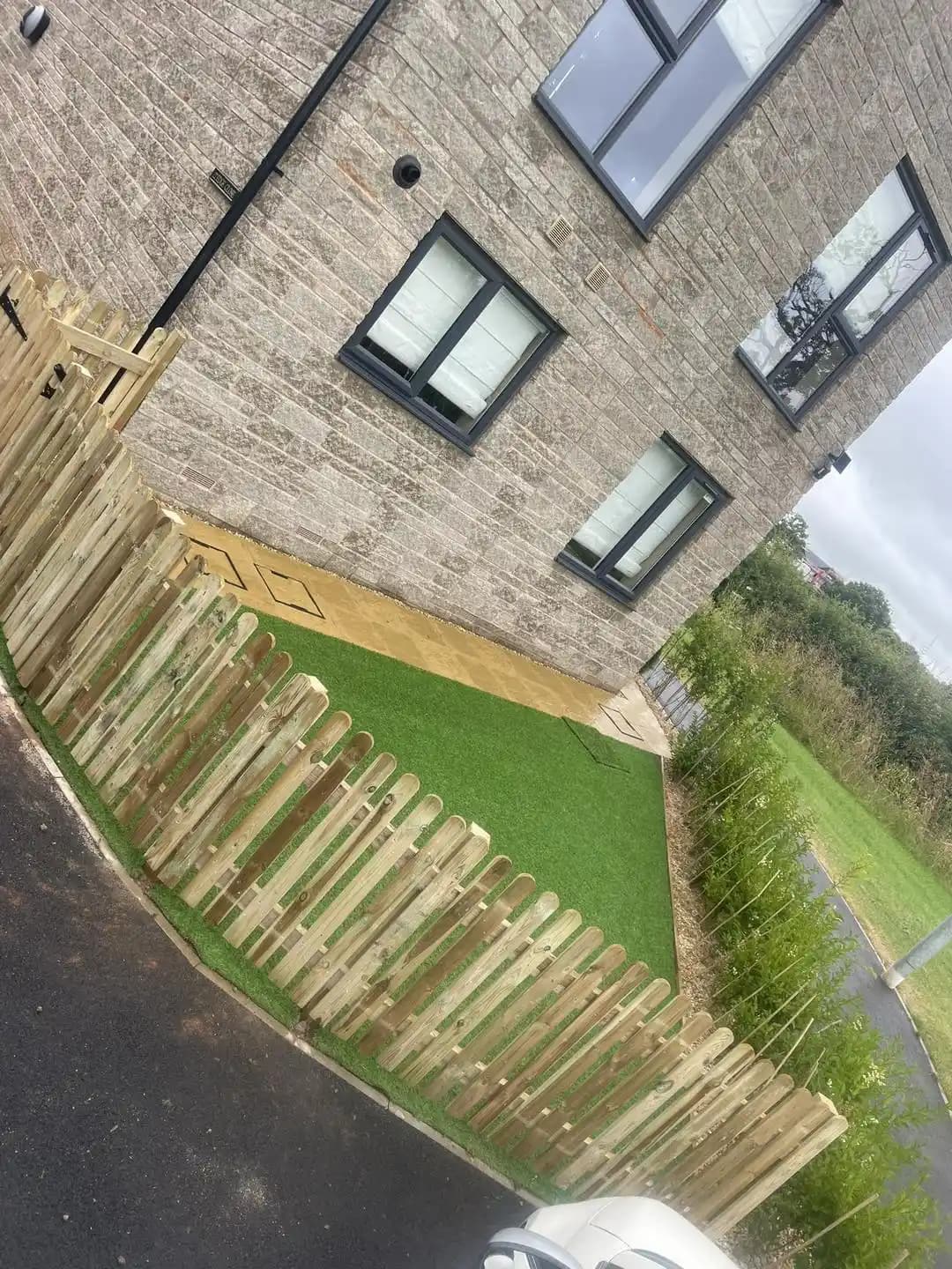 Small side garden with artificial grass enclosed by a low timber picket fence beside a modern house.