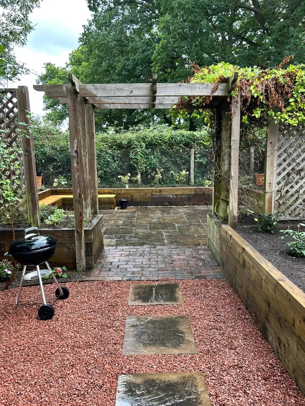 Black metal garden arch set between trellis panels with paving and planted borders in a sunny garden.
