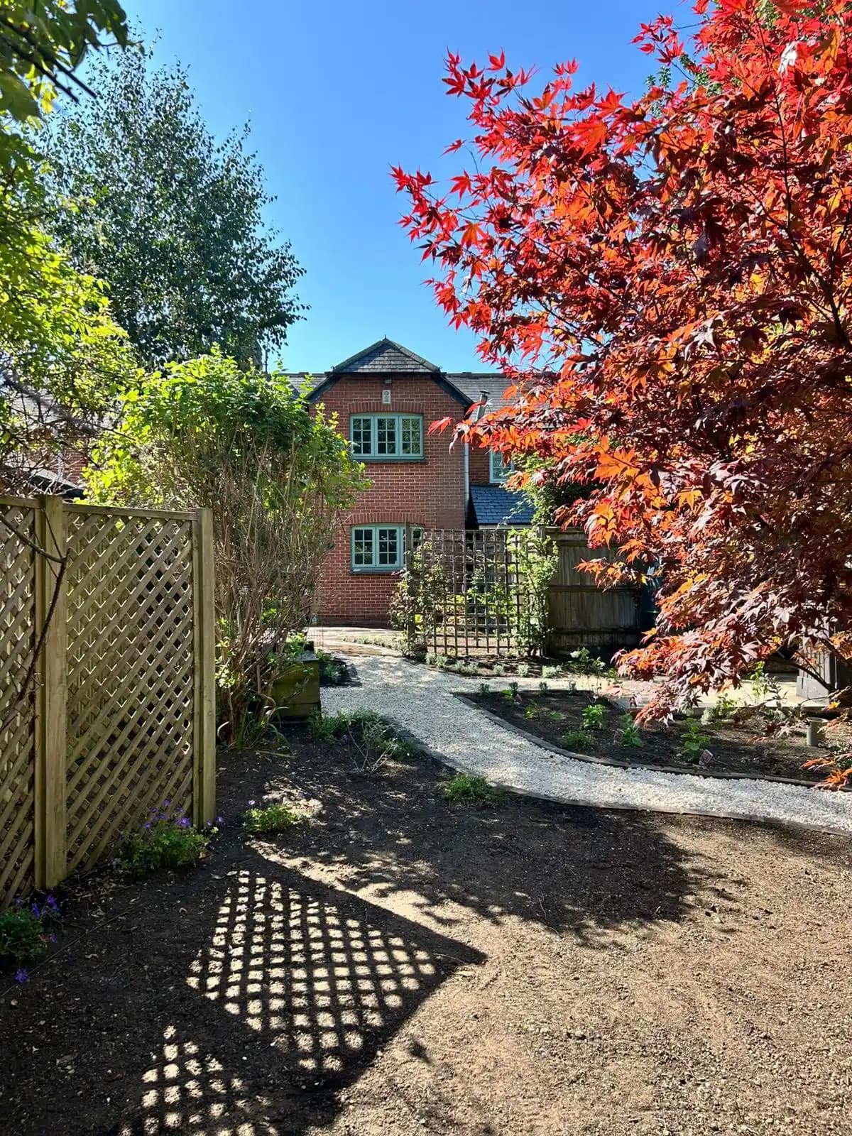 Curved gravel garden path framed by trellis panels, planting beds and a red Japanese acer tree.