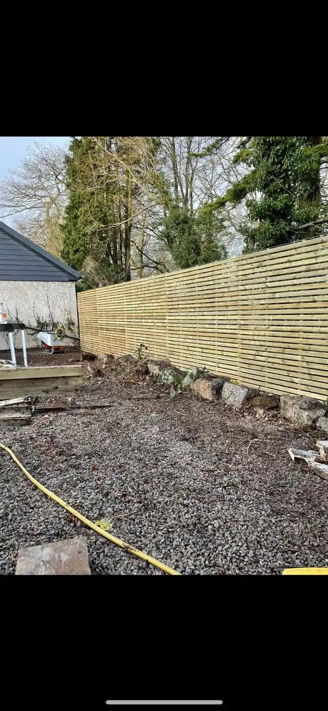 Horizontal slatted timber privacy fence installed along a gravel garden boundary beneath mature trees.