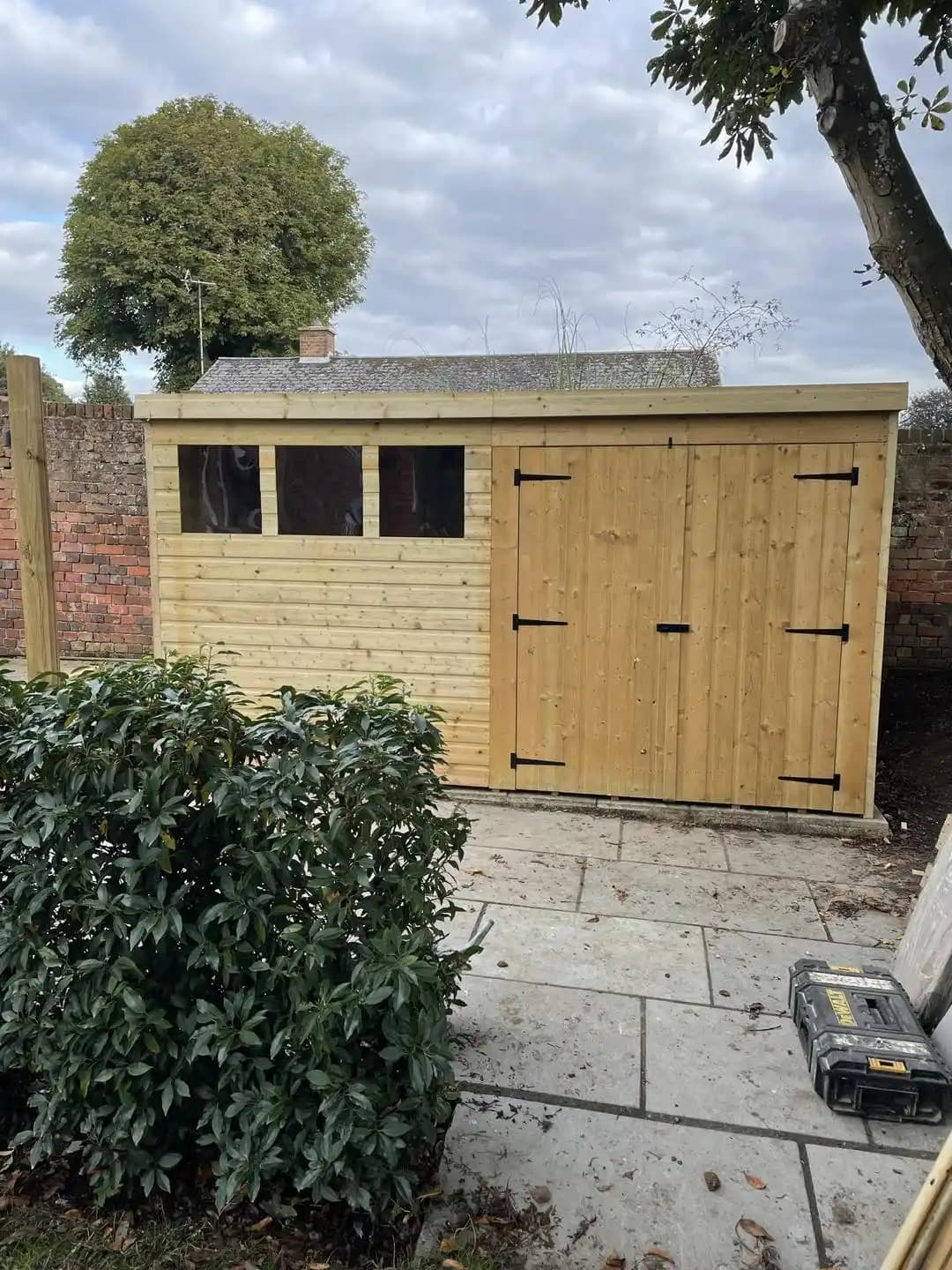 Large timber garden shed with double doors and windows installed on a paved patio area.