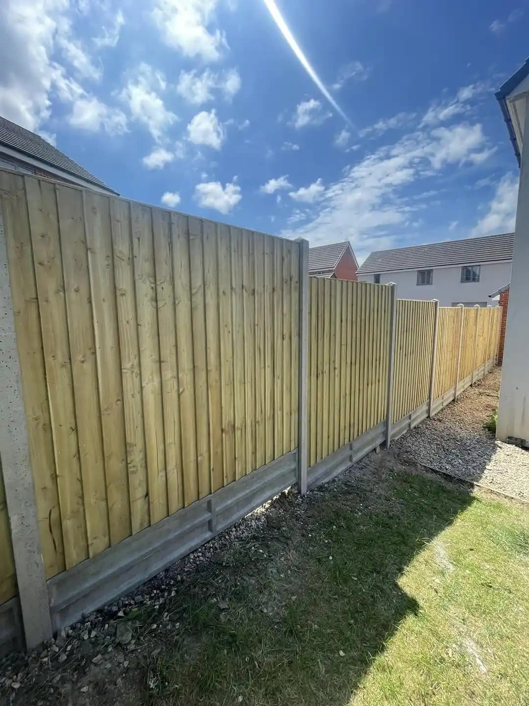 Long closeboard boundary fence with concrete posts running beside a house under a blue sky.