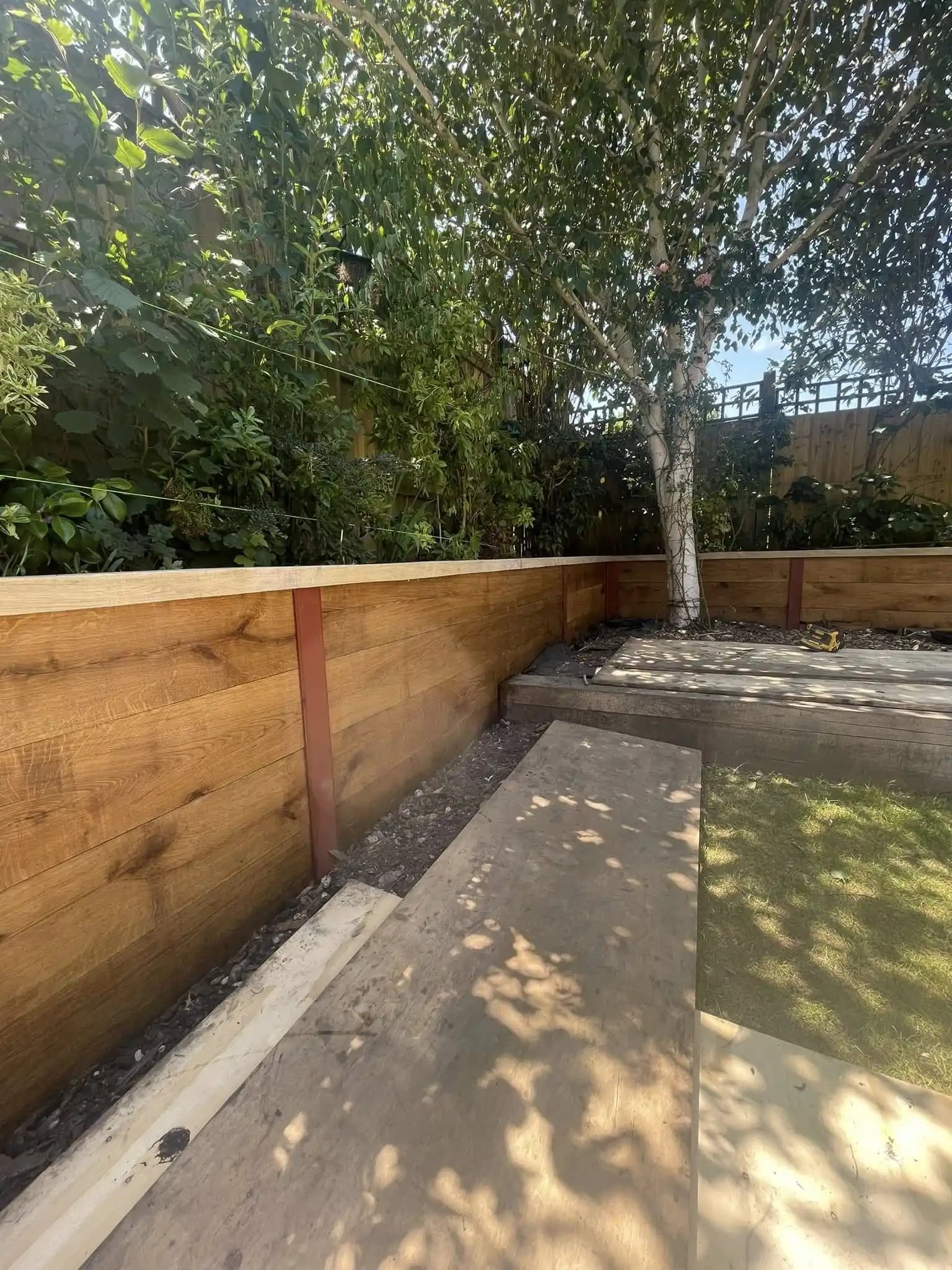 New oak-style sleeper retaining wall with steel posts around a garden border and tree during landscaping work.