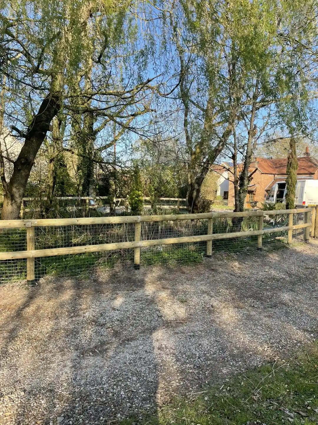Long run of post and rail fencing with wire mesh beside a gravel driveway and trees in a rural setting.