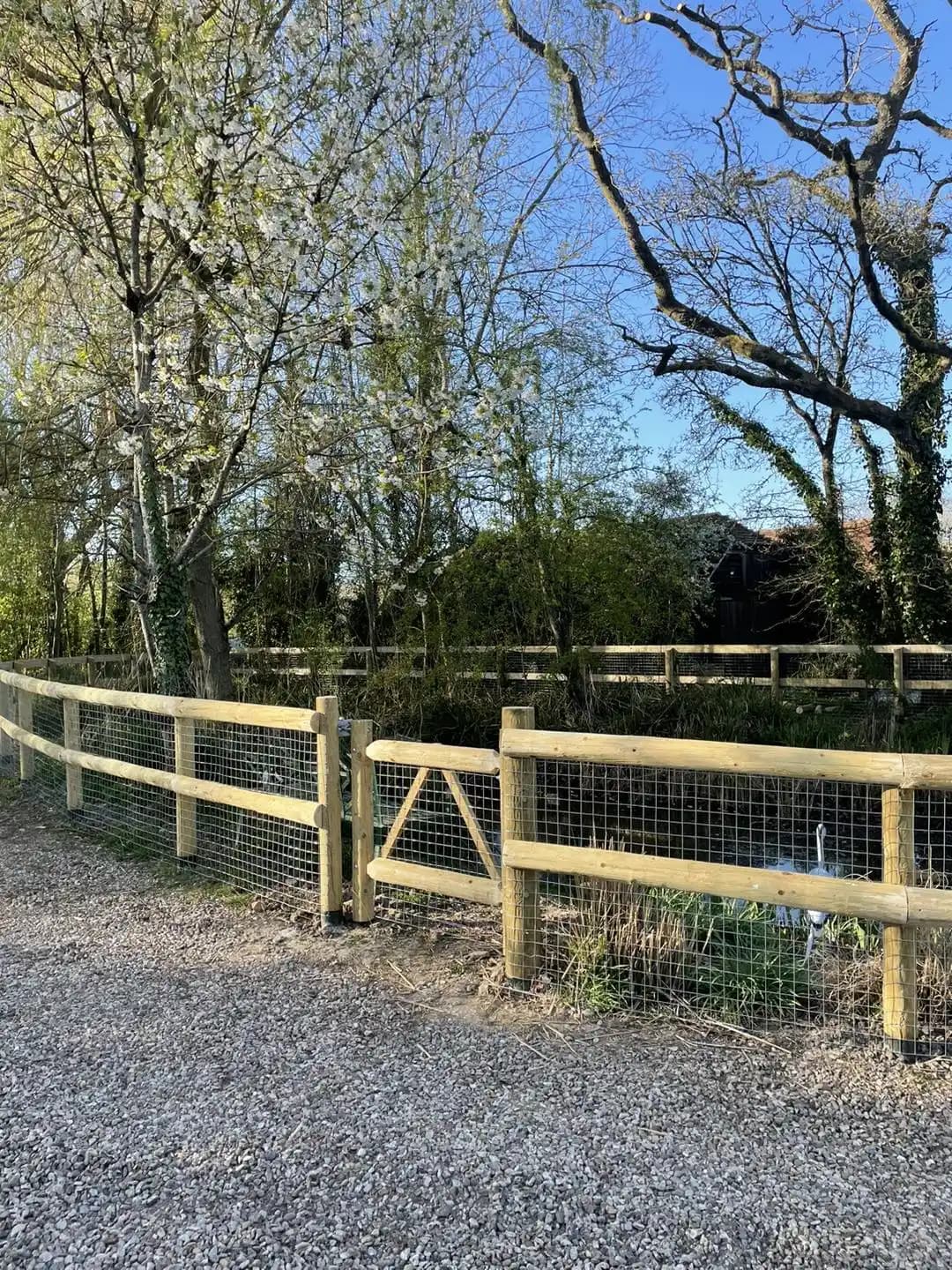 Timber post and rail fencing with wire mesh and gate creating a secure boundary beside a gravel drive.