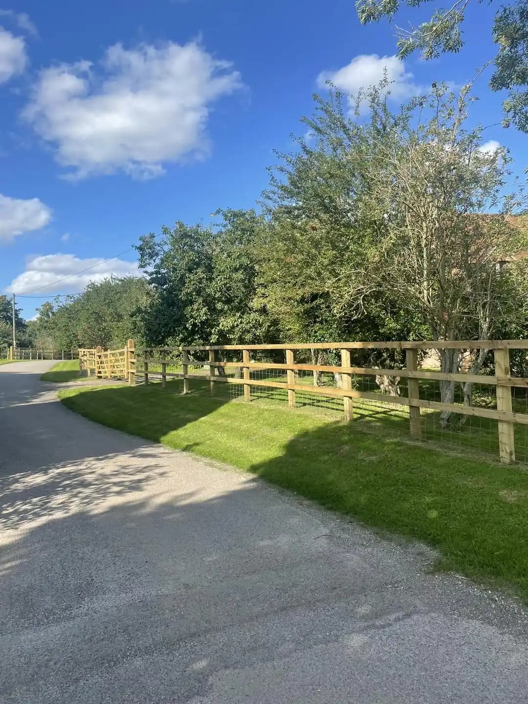 Post and rail timber fence with wire mesh running alongside a country lane and grass verge.