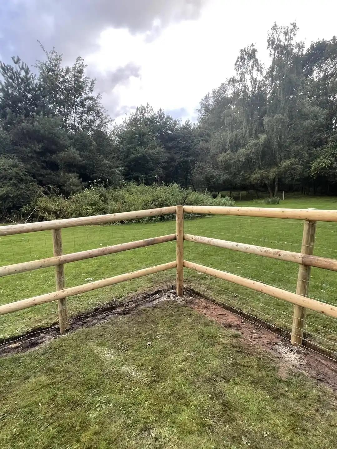 Corner of a post and rail fence with wire mesh enclosing a lawn and garden boundary.