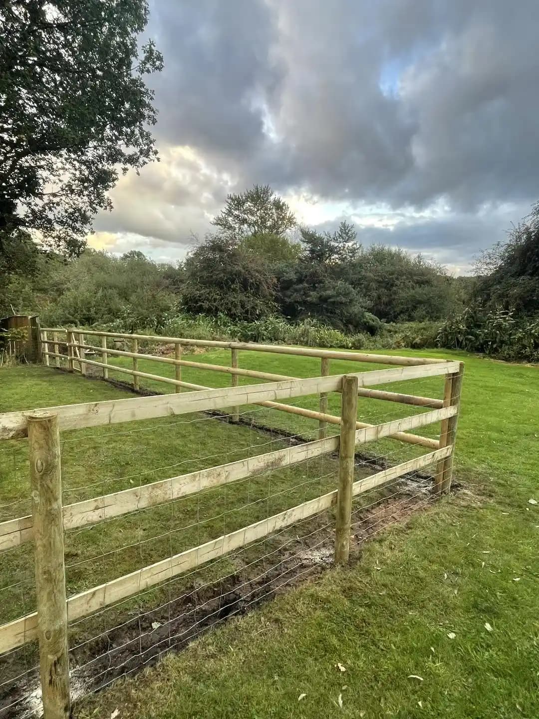 Post and rail timber fence with wire mesh enclosing a rural garden boundary with mature trees beyond.