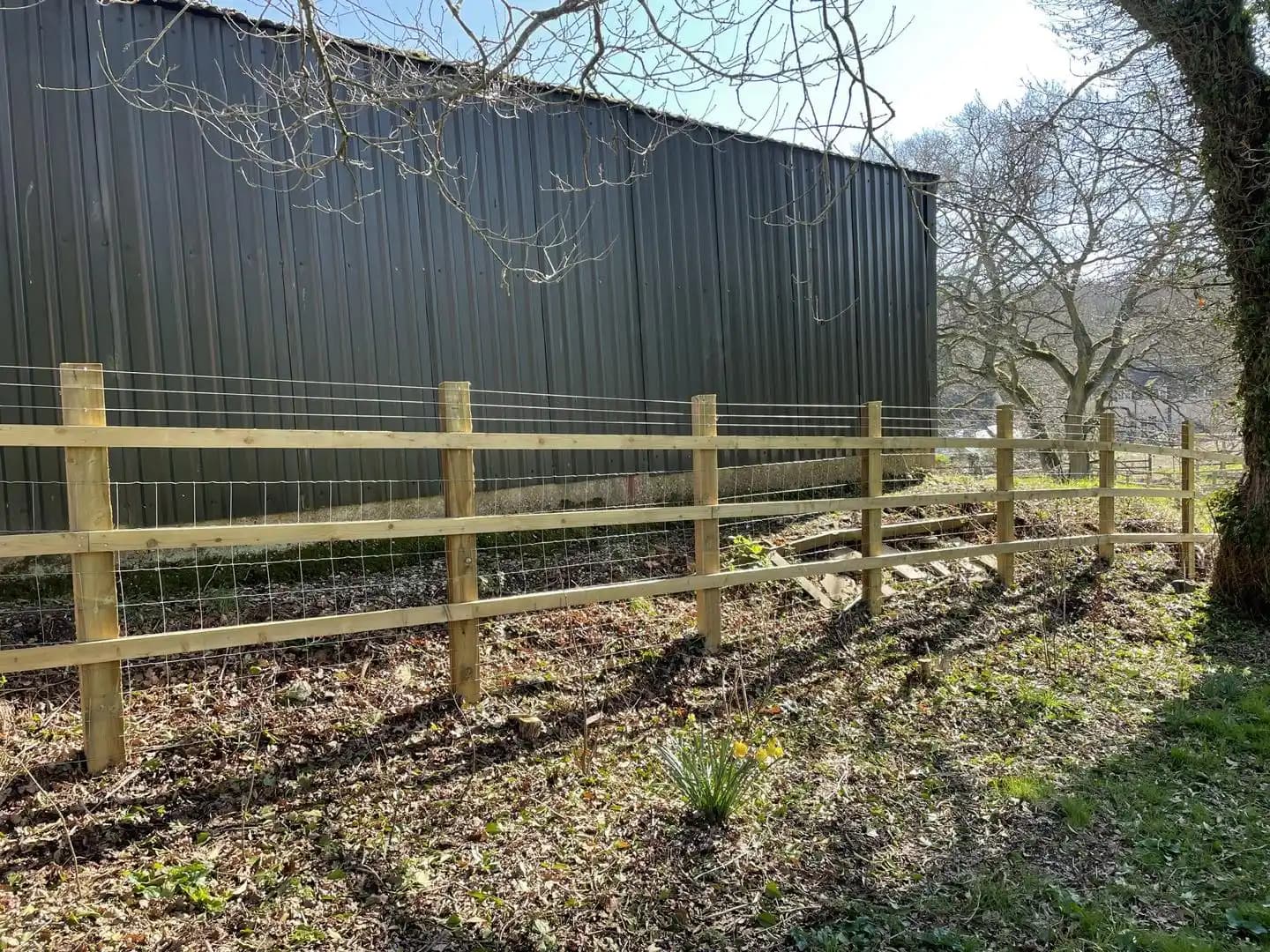 Post and rail fence with wire mesh installed beside a black outbuilding and wooded boundary.