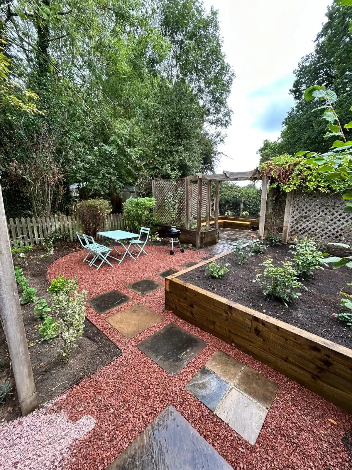 Red gravel seating area with stepping stones, raised sleeper bed, pergola and trellis screens in a finished garden.