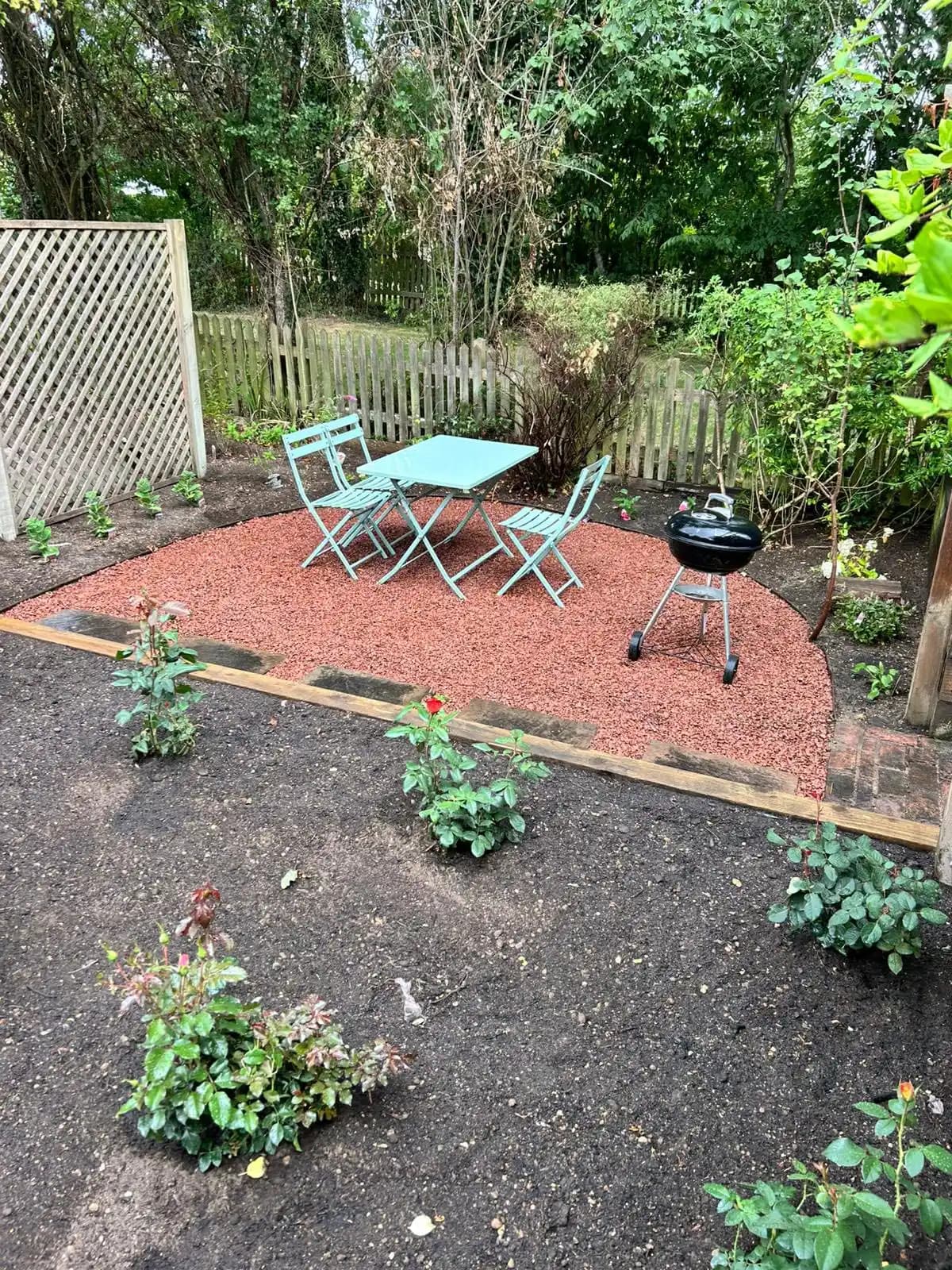 New red gravel seating area with bistro table, barbecue, planted borders and timber edging in a landscaped garden.