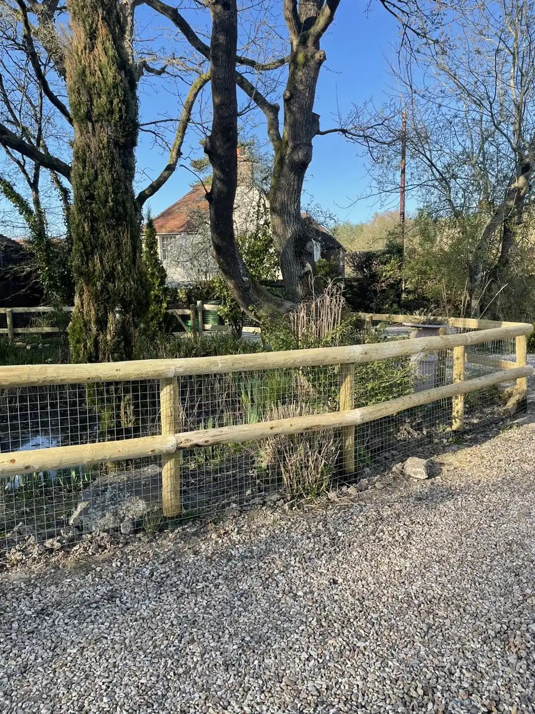 Curved run of post and rail fencing with wire mesh following a gravel drive through mature trees.