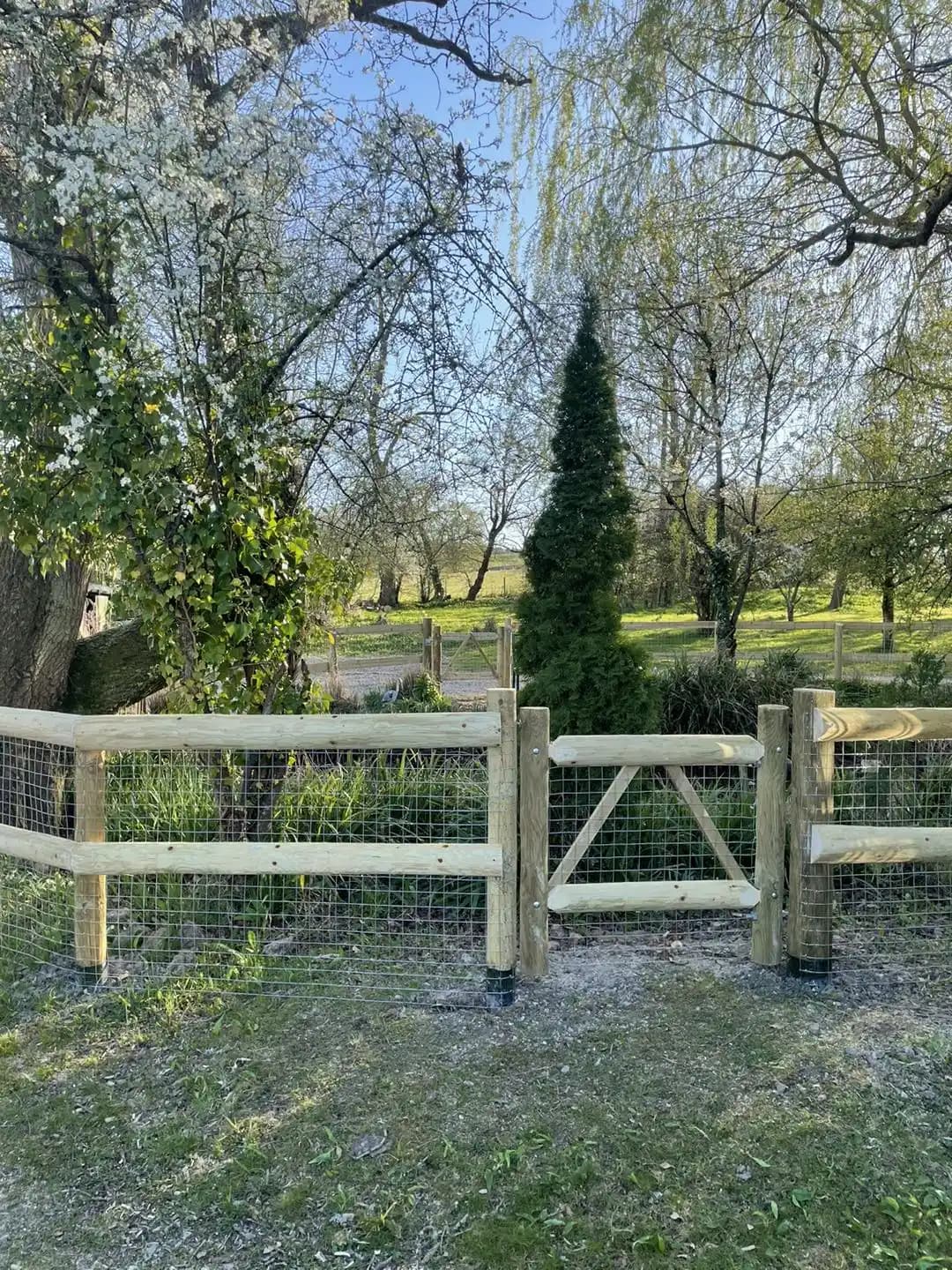 New post and rail wire mesh fence with matching timber gate enclosing a rural garden boundary.