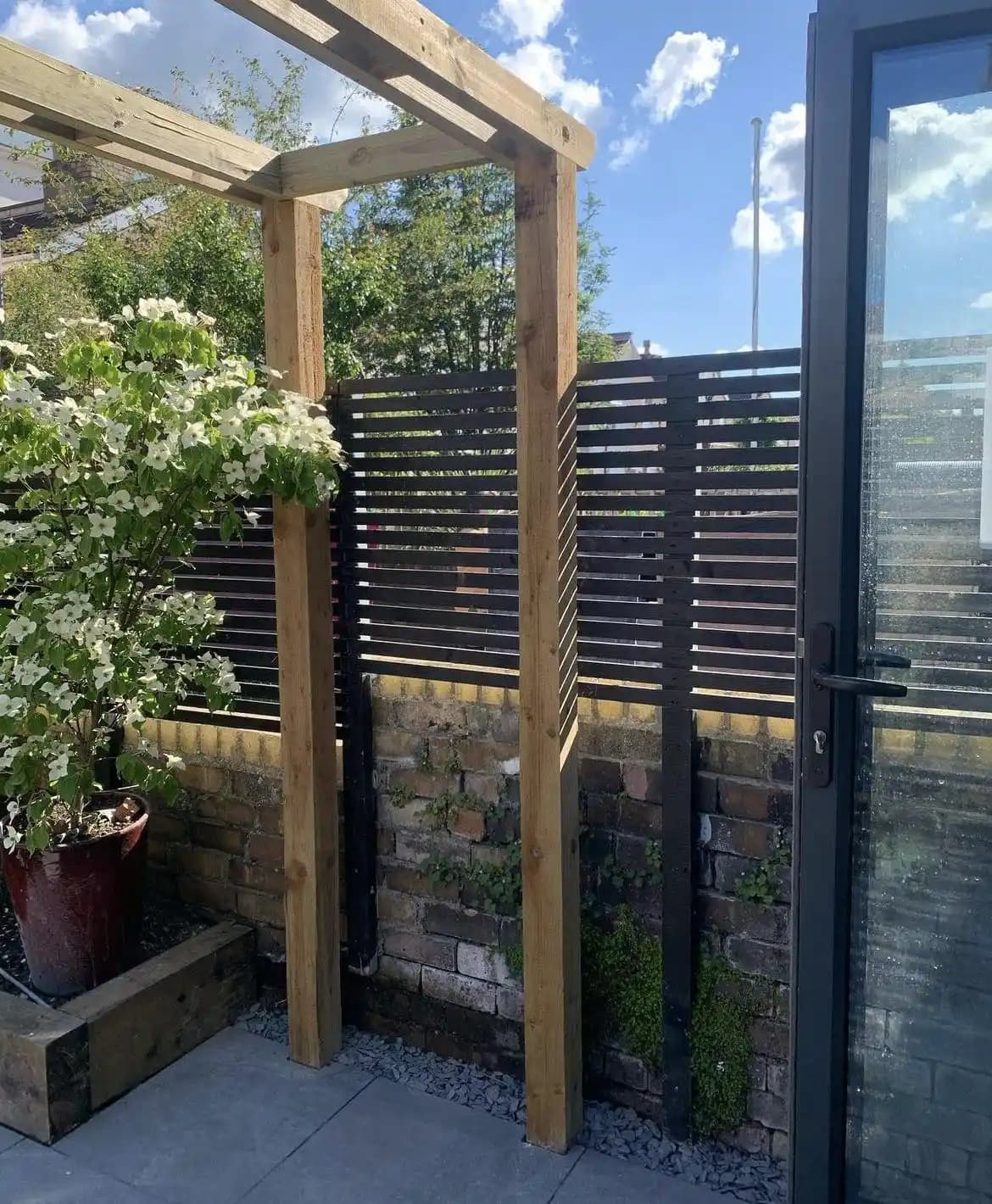 Timber pergola posts installed beside a black horizontal slatted privacy screen above a brick wall.