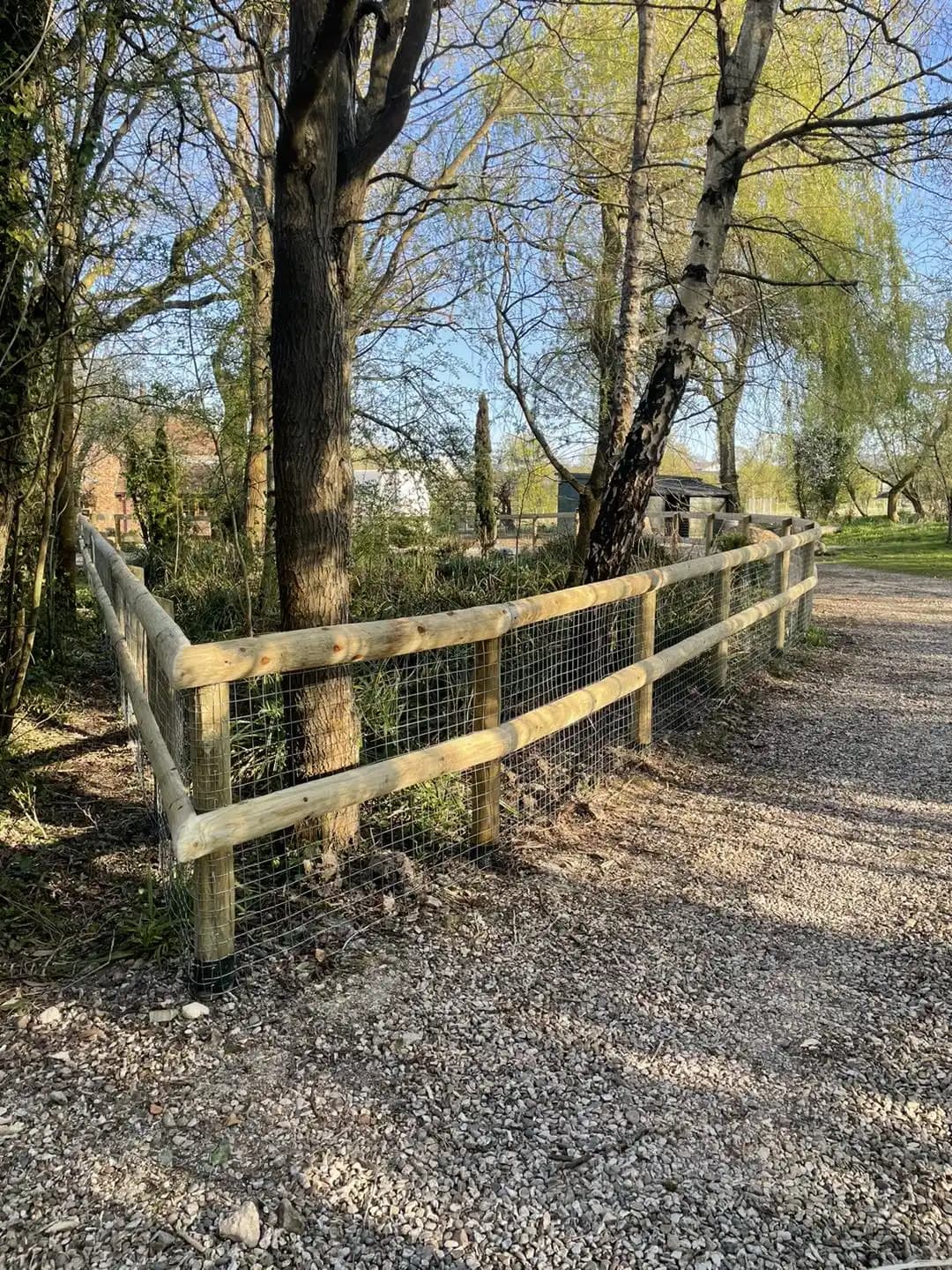 Post and rail boundary fence with wire mesh installed around mature trees and a gravel access area.
